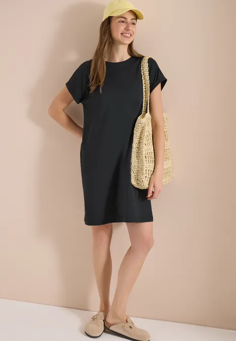 Frau in lockerem schwarzen T-Shirt-Kleid mit Basttasche und beigen Slip-on-Schuhen.