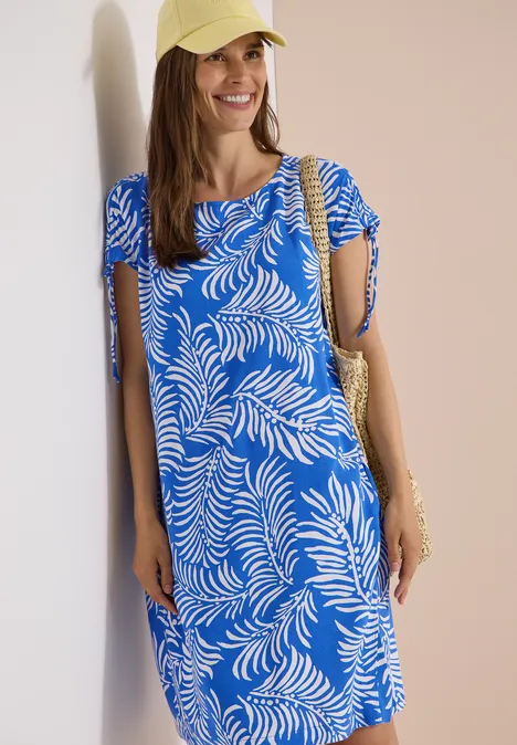 Frau in blau-weiß gemustertem Kleid, Strohhut und Tasche steht an einer Wand.