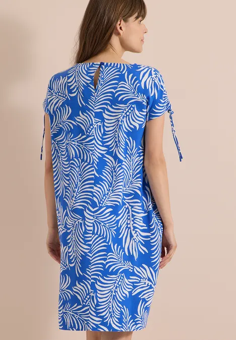 Frau von hinten in knielangem, blauem Kleid mit weißem Blattmuster, kurzen Ärmeln und seitlichen Schleifen.