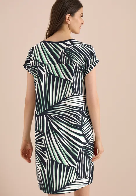 Knielanges Kleid aus Jersey palm breeze green Frau von hinten in kurzärmeligem Kleid mit blau-grünem Palmenmuster vor beigem Hintergrund.