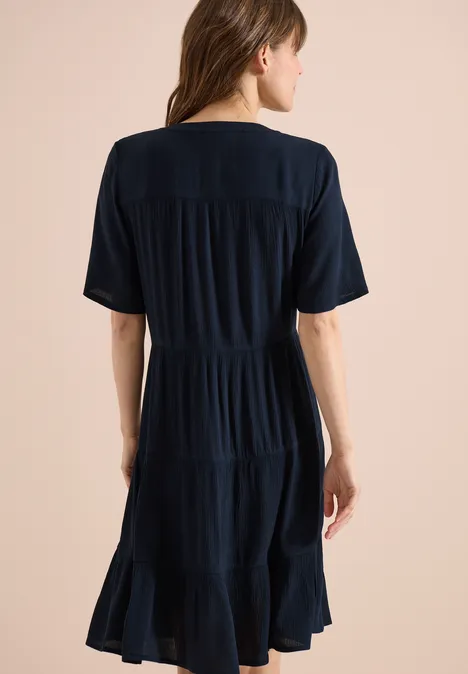 Rückenansicht einer Frau in einem knielangen, dunkelblauen, kurzärmeligen Kleid mit stufigem Rocksaum.