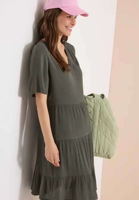 Frau in khakigrünem Stufenkleid mit kurzen Ärmeln, hält eine gesteppte Umhängetasche.