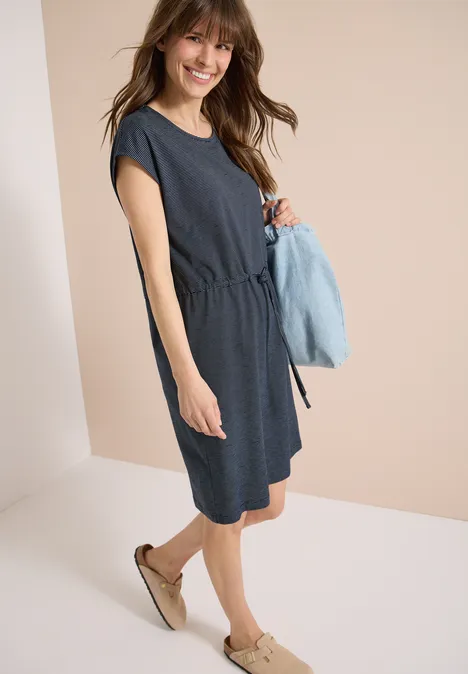 Knielanges Kleid aus Jersey universal blue Frau in knielangem, dunkelblauem Freizeitkleid mit kurzen Ärmeln und Espadrilles, hält hellblauen Pullover.