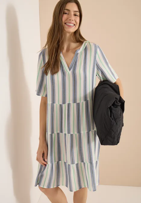 Vrouw in midi-jurk met pastelkleurige strepen, houdend een zwarte jas tegen een neutrale achtergrond.