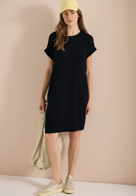 Vrouw in zwarte T-shirtjurk, sneakers met tas, voor een neutrale achtergrond.