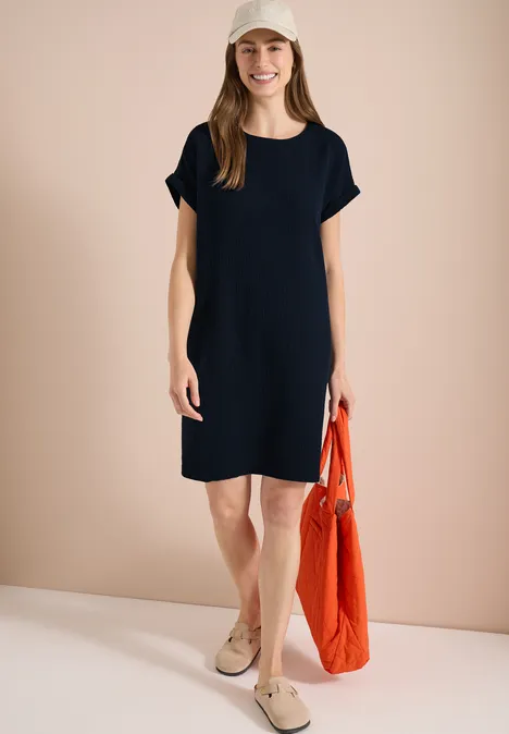 Femme en robe noire à manches courtes, chaussures beiges et sac orange.