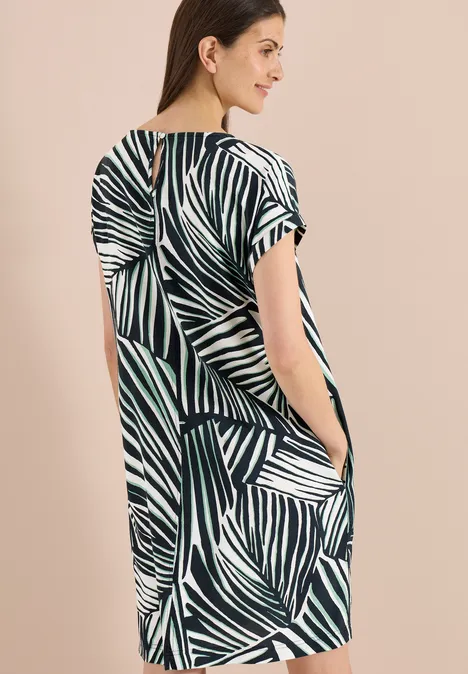 Frau vor hellem Hintergrund in locker fallendem Kurzarmkleid mit grün-weißem Blattmuster und seitlichen Taschen.