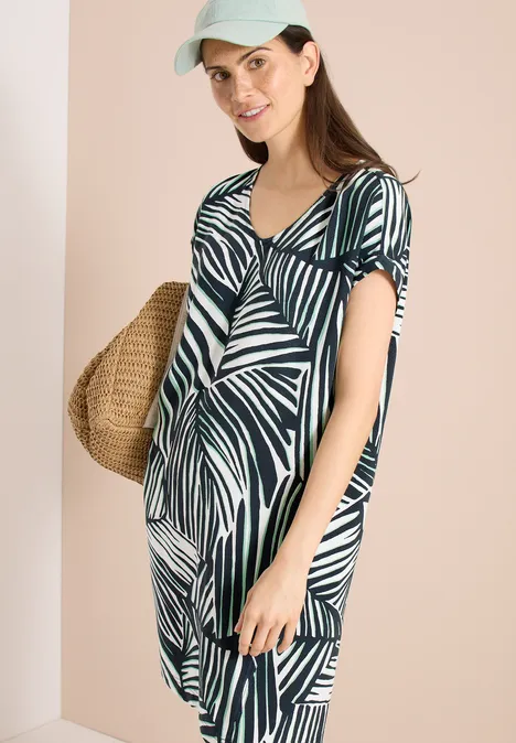 Frau in sommerlichem grün-weißem Blattmuster-Kleid mit Korbtasche vor beigem Hintergrund.