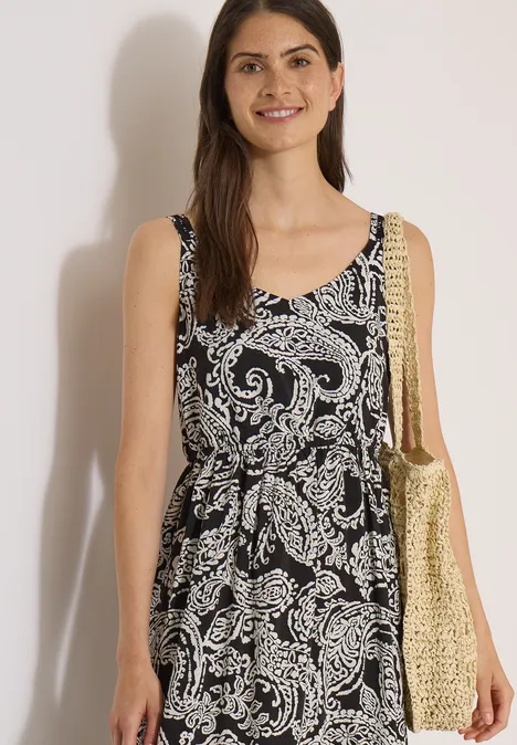 Frau trägt ärmelloses schwarzes Sommerkleid mit weißem Paisley-Muster und goldfarbener Stroh-Tasche.