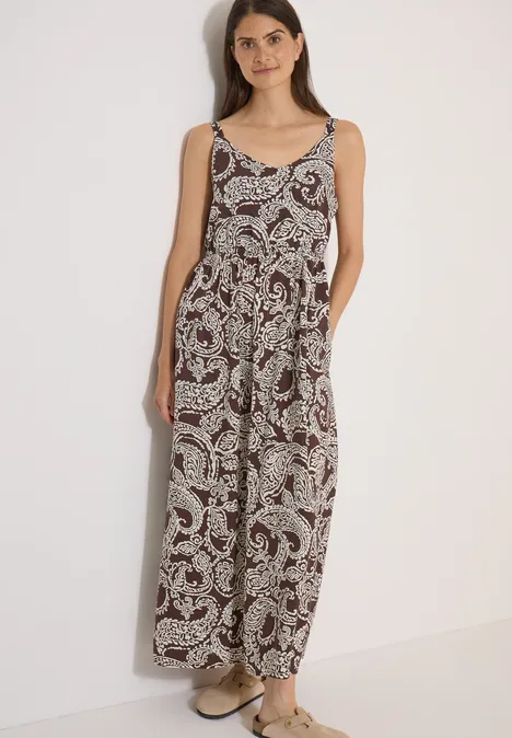 Robe longue à encolure en V et imprimé brown cacao Femme en combinaison ample sans manches à motif paisley et sandales devant un mur clair.