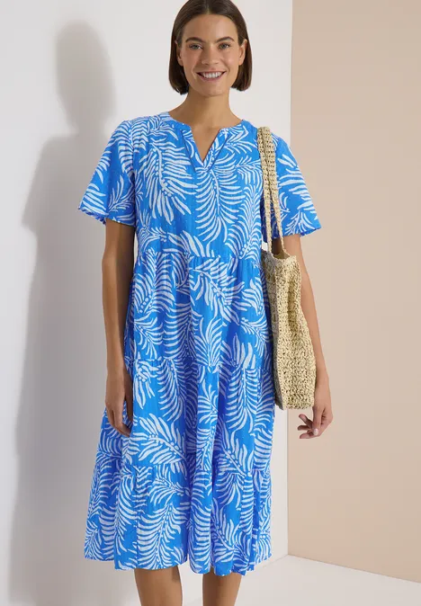 Midikleid mit Split Neck und Print ethereal blue Frau in blauem Midikleid mit weißem Blattmuster, trägt eine geflochtene Strohtasche an der Schulter.