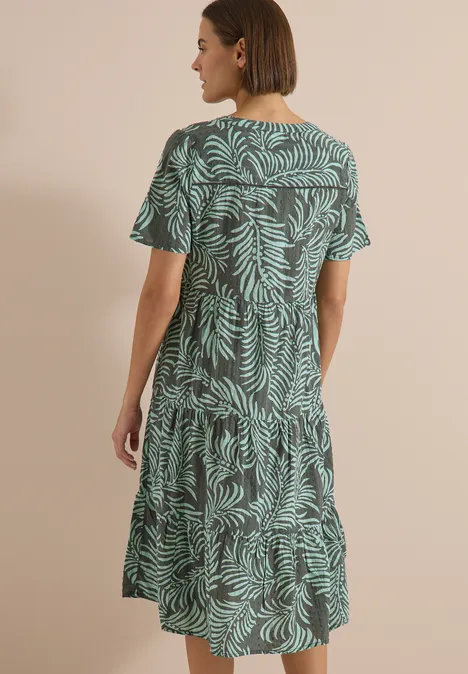 Midikleid mit Split Neck und Print khaki Eine Frau steht mit dem Rücken zur Kamera, trägt ein knielanges, locker sitzendes Kleid mit grünem Blattmuster.