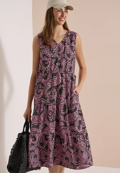 Frau in ärmellosem, lila gemustertem Midikleid mit Taschen, schwarzer Flecht-Tasche und beiger Kappe.