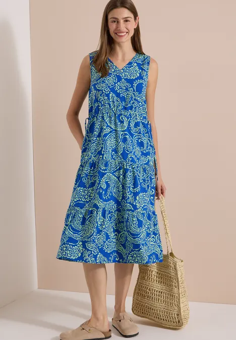 Midijurk met V-hals en print surf blue Vrouw in mouwloze midi-jurk met blauw dessin, draagt een strooien tas en comfortabele sandalen.
