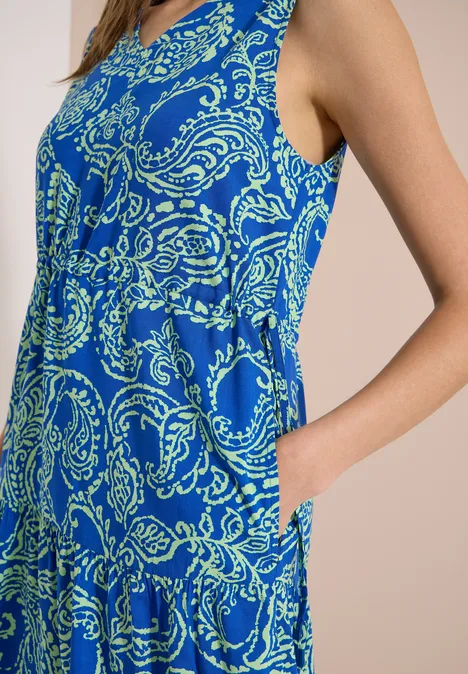 Midijurk met V-hals en print surf blue Vrouw in zijaanzicht met een mouwloze, blauwe jurk met paisleyprint, hand in de zak.