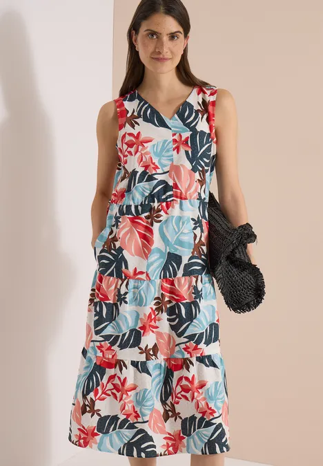 Vrouw in mouwloze zomerse jurk met lagen en kleurrijk bloemen- en bladmotief, met een zwarte gehaakte tas.