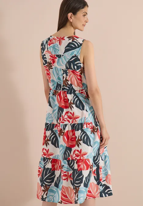 Vrouw van achteren in een mouwloze jurk met kleurrijk bloemen- en bladmotief tegen een beige achtergrond.