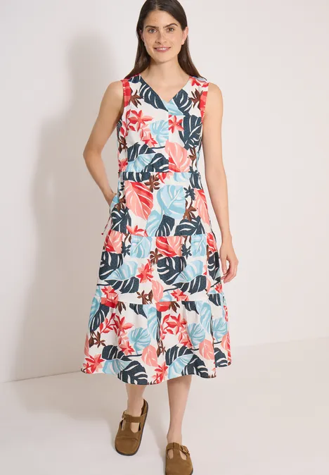 Vrouw in mouwloze midi-jurk wit met rood-blauw tropisch bloemenbladmotief, bruine schoenen, voor een lichte achtergrond.