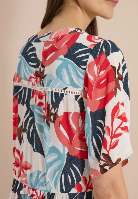 Robe mi-longue à col fendu et imprimé light alabaster white Femme vue de dos portant un haut blanc à manches mi-longues avec un motif feuilles tropicales rouge et bleu.
