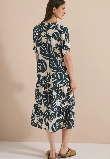 Femme de dos en robe mi-longue beige à imprimé feuilles bleues, sandales.