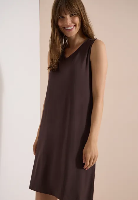Frau trägt ärmelloses, knielanges, braunes Kleid mit schlichter Silhouette.