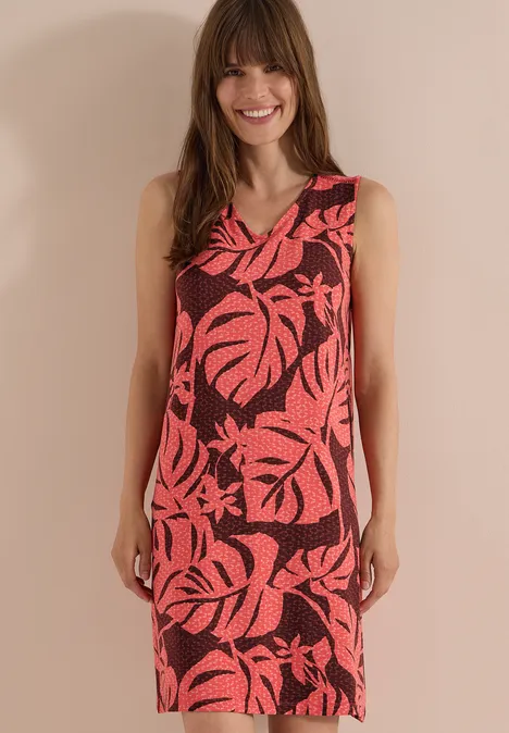 Knielanges Jerseykleid mit V-Neckund Print coral Frau in ärmellosem roten Kleid mit schwarzem Tropenblattmuster vor beigem Hintergrund.