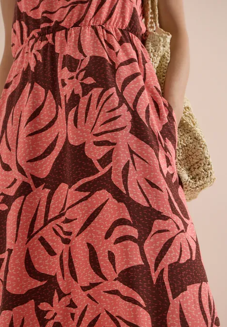 Robe longue rouge à imprimé feuilles tropicales, femme les mains dans les poches, avec sac en paille.