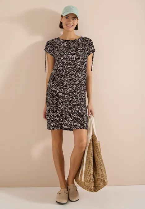Robe mi-longue avec manchons brown cacao Femme en robe courte noire et blanche à pois, chapeau, mules et sac en paille sur fond beige.