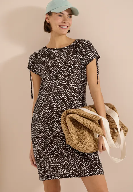 Robe mi-longue avec manchons brown cacao Femme en robe à manches courtes à motifs, portant un sac de plage en paille et une casquette.