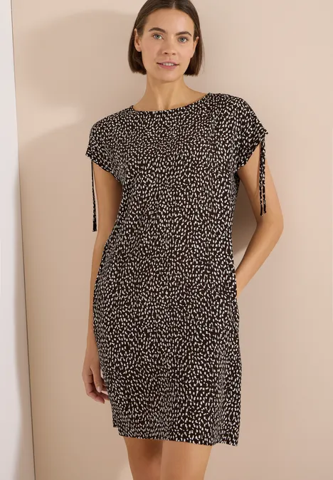Robe mi-longue avec manchons brown cacao Une femme porte une robe noire aux petits pois blancs jusqu’aux genoux avec manches courtes.