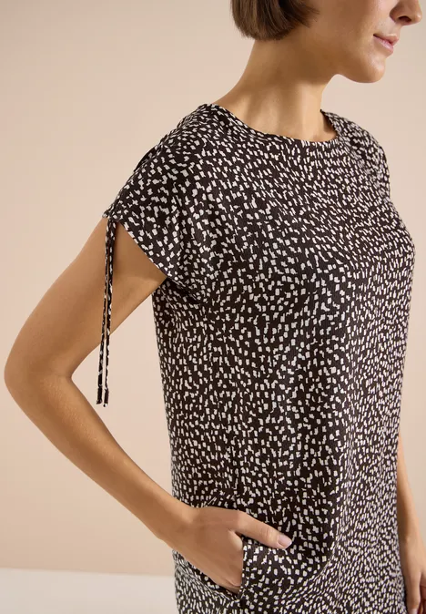Robe mi-longue avec manchons brown cacao Femme portant un haut noir à motifs blancs, manches courtes avec lien latéral, main dans la poche.