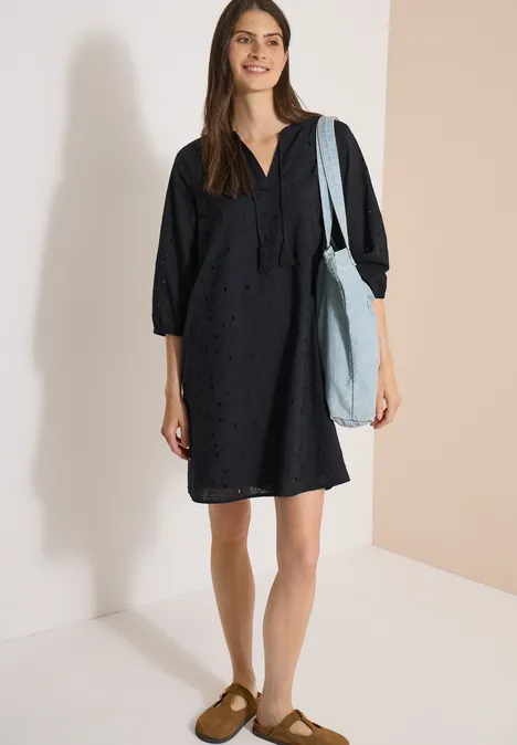 Robe longueur genou avec manches 3/4 et broderie Black Femme portant une robe noire ample, un sac bleu clair à l'épaule et des mocassins marron sur fond neutre.