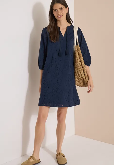 Robe longueur genou avec manches 3/4 et broderie universal blue Femme en robe courte bleu marine en dentelle à capuche, portant un sac en osier et des chaussures plates.
