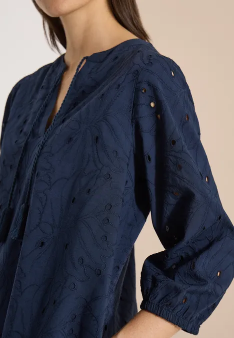 Robe longueur genou avec manches 3/4 et broderie universal blue Haut féminin bleu marine brodé avec ajours et manches longues, posé devant un fond clair.