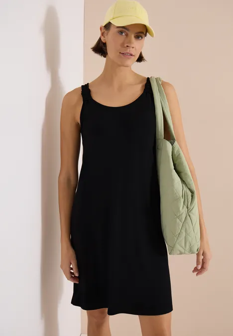 Person in ärmellosem schwarzem Kleid, gesteppter grüner Tasche und heller Mütze vor hellem Hintergrund.