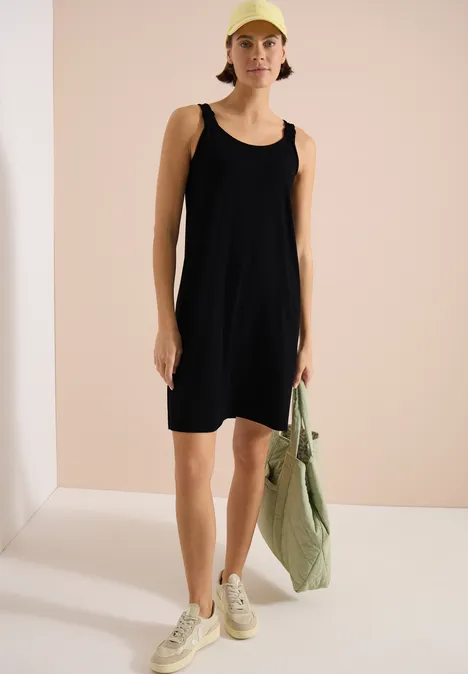 Schwarzes ärmelloses Kleid, helle Sneaker, grün-gestreifte Tasche, schlichter Raum.