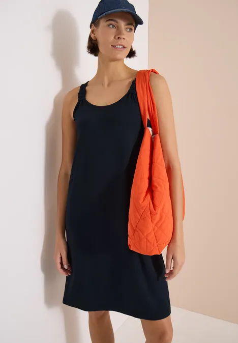 Person trägt ärmelloses schwarzes Kleid, orange gesteppte Tasche und Baseballcap vor neutralem Hintergrund.