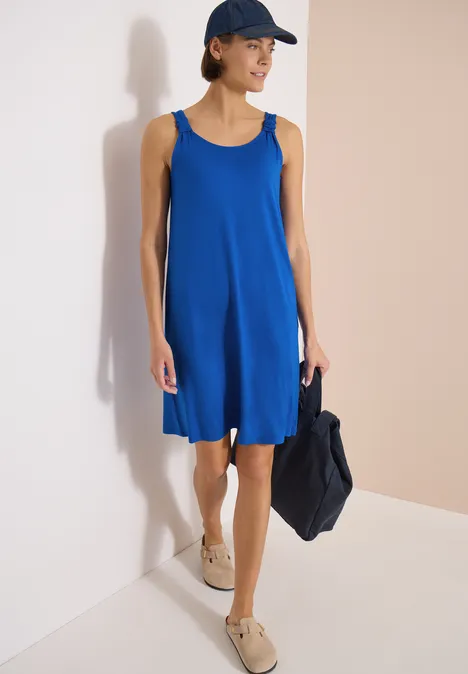 Vrouw in losvallende, mouwloze blauwe jurk met pet, draagt een donkerblauwe tas en beige espadrilles.