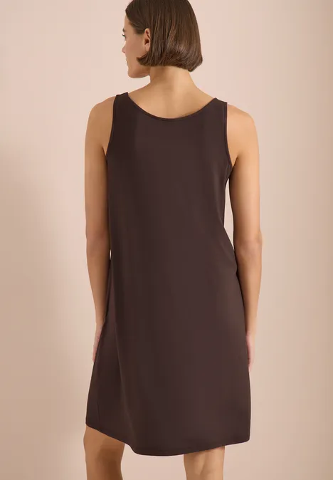 Eine Frau von hinten in einem ärmellosen braunen Kleid vor hellem Hintergrund.