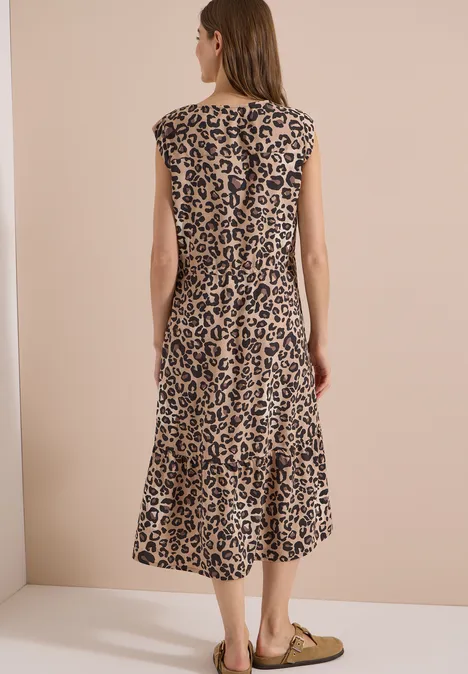 Frau von hinten in ärmellosem, midilangen Leopardenkleid und flachen Ballerinas vor beigem Hintergrund.