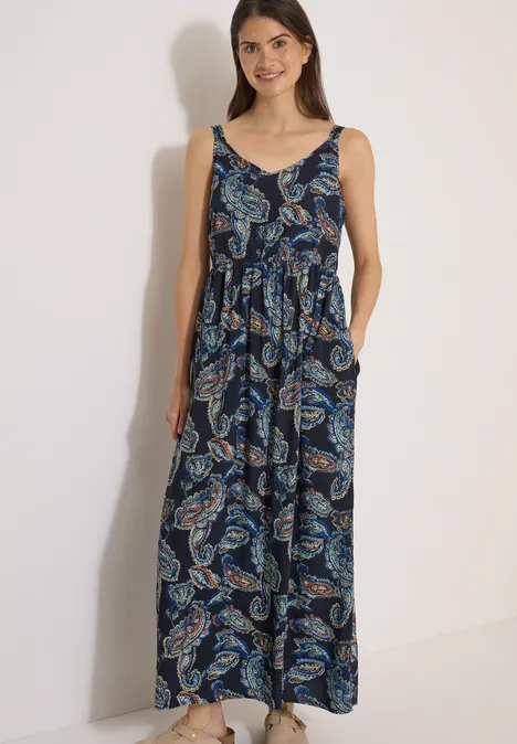 Frau trägt ärmelloses, bodenlanges Maxikleid mit Paisley-Muster in Blau- und Grüntönen.