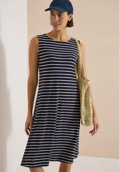 Frau in blau-weiß gestreiftem Midi-Kleid mit Strohtasche und Cap vor hellem Hintergrund.