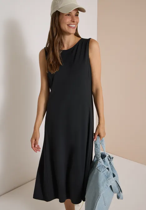 Frau in schwarzem ärmellosem Midikleid mit Cap hält blaue Jeansjacke und lächelt.