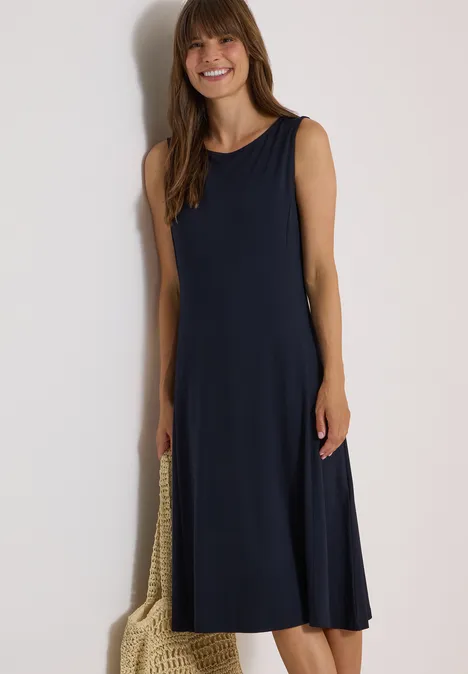 Vrouw in mouwloze donkerblauwe midi-jurk met nettas, glimlachend.