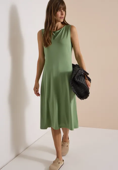 Frau in ärmellosem grünen Midikleid, trägt Espadrilles und schwarze Clutch, vor neutralem Hintergrund.