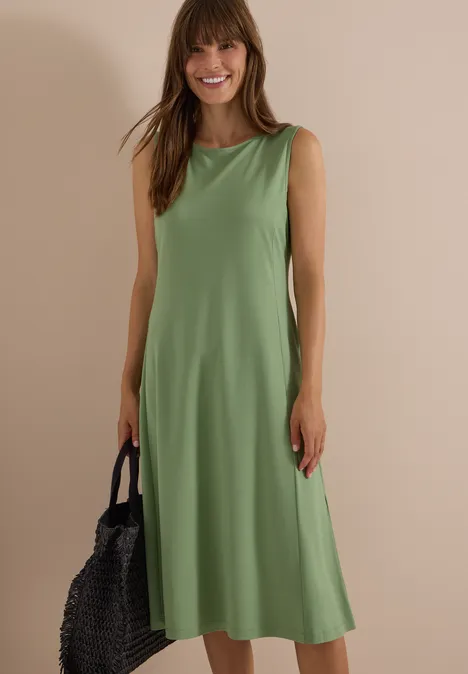 Frau in ärmellosem, grünem Midi-Kleid, lächelnd, hält schwarze, geflochtene Tasche, neutraler Hintergrund.