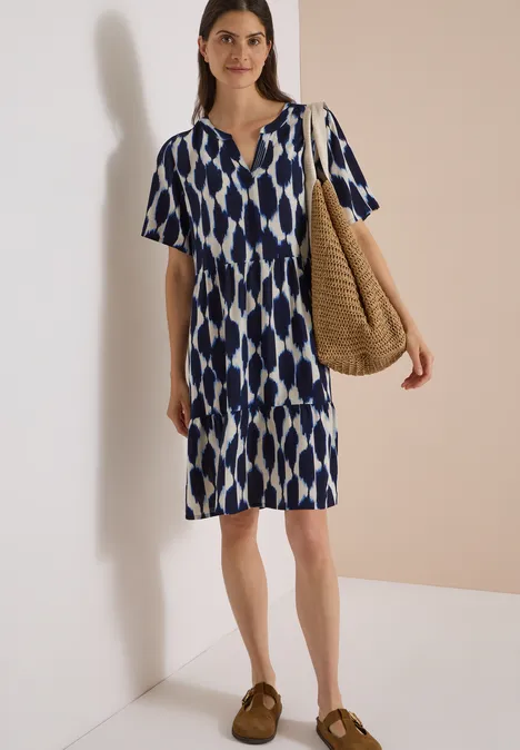 Knielanges Kleid mit Split Neck und Struktur urban dark blue Frau trägt knielanges blau-weißes buntes Musterkleid mit kurzen Ärmeln und Basttasche.