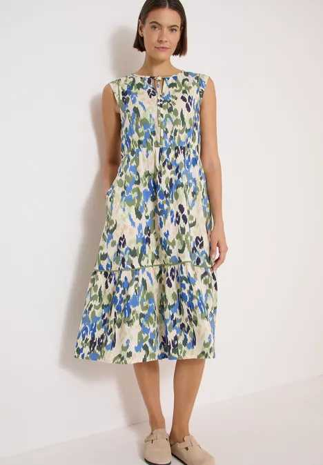 Frau steht vor weißem Hintergrund, trägt ärmelloses Midikleid mit grün-blauem Blumenmuster und Stufenrock.
