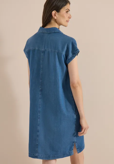 Vrouw van achteren in een losse jeansoverhemdjurk met korte mouwen voor een neutrale achtergrond.