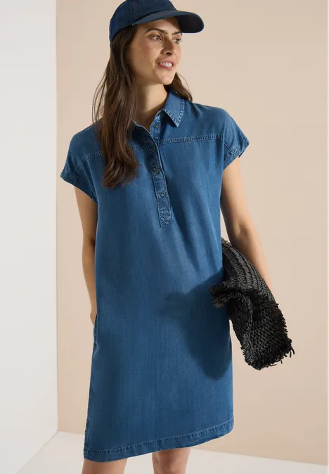 Vrouw in blauwe denimjurk met pet, draagt zwarte tas voor lichte achtergrond.